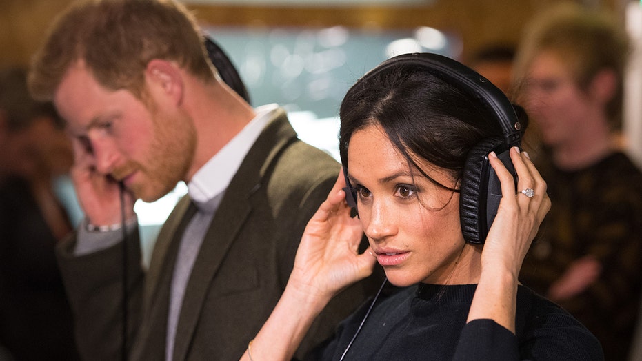 A close-up of Meghan Markle wearing a black sweater listening from headphones
