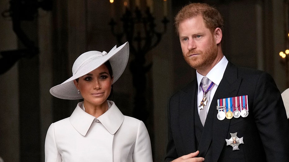 Meghan Markle wearing a white coat dress with a matching hat standing next to Prince Harri in a suit and tie